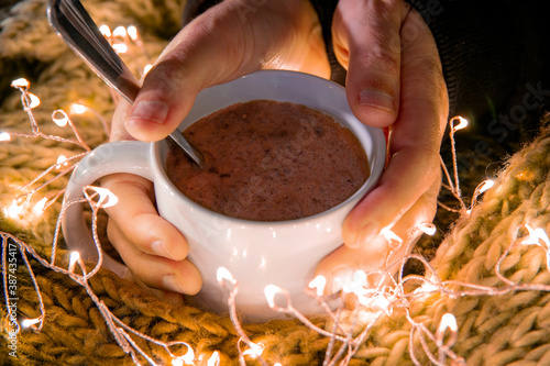 Close-up of hands holding a bowl of hot chocolate between a woolen blanket and Christmas lights. Winter concept at home. Horizontal photography. Christmas time