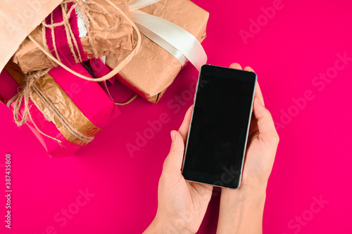 Cropped view of woman holding smartphone with blank screen on pink background with christmas gifts.