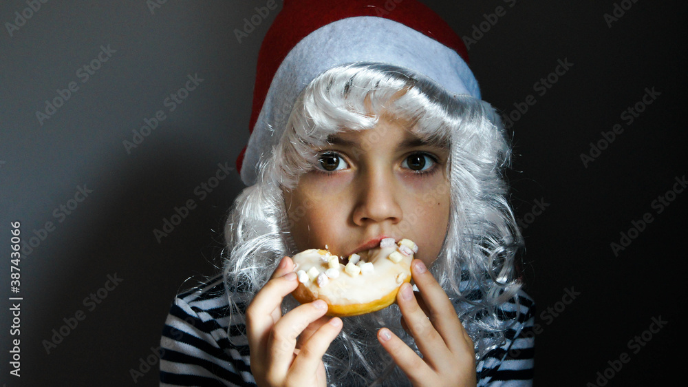 Serious boy in a Santa Claus costume is eating tasty doughnut in the grey background. New year and Merry Christmas fast food and bakery concept Serious boy in a Santa Claus costume is eating tasty doughnut in the grey background. New year and Merry Christmas fast food and bakery concept