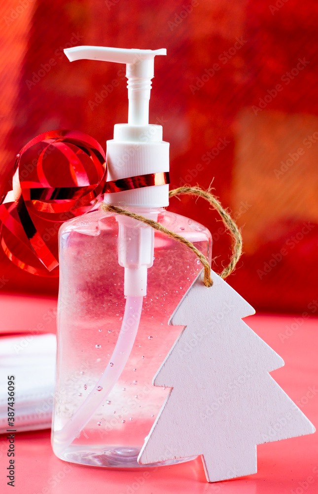 Bottle of hand gel sanitizer with Christmas decoration and ribbon as a gift. Concept of spending Christmas during coronavirus pandemic lock down and quarantine. Red glittery background.