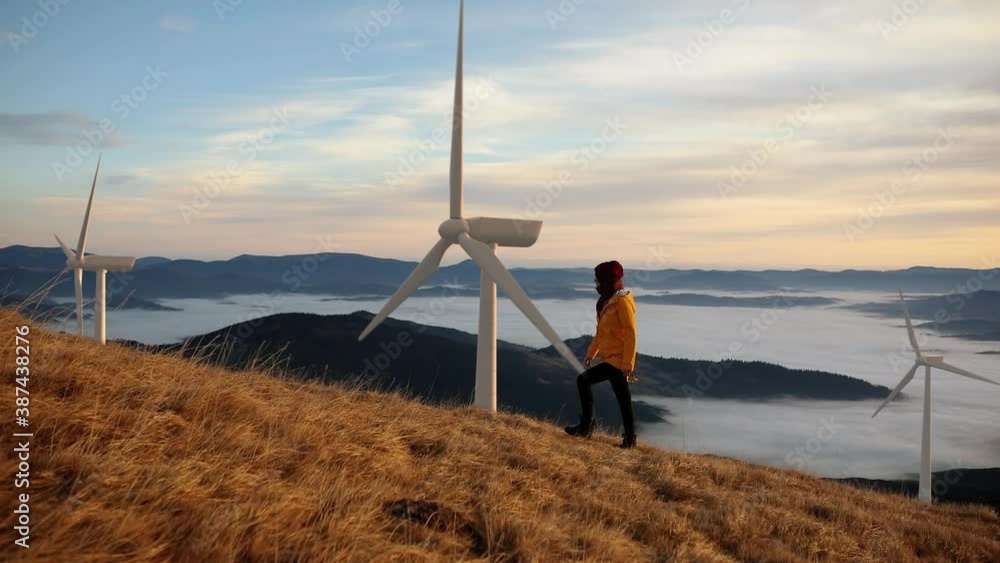 Epic shot of a woman hiking on the edge of the mountain against landscape with wind turbine power station on background. Concept of: environmental engineering, renewable energy and love for nature