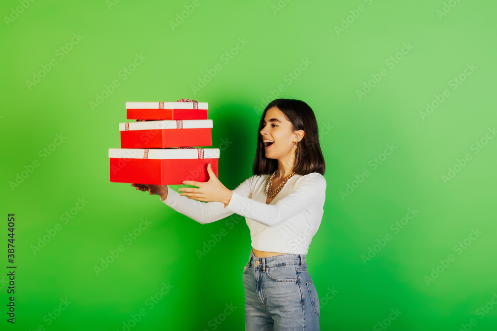 Happy young brunette girl in casual clothes is throwing up red-white present boxes keeping mouth open isolated on green background. Happy young brunette girl in casual clothes is throwing up red-white present boxes keeping mouth open isolated on green background.