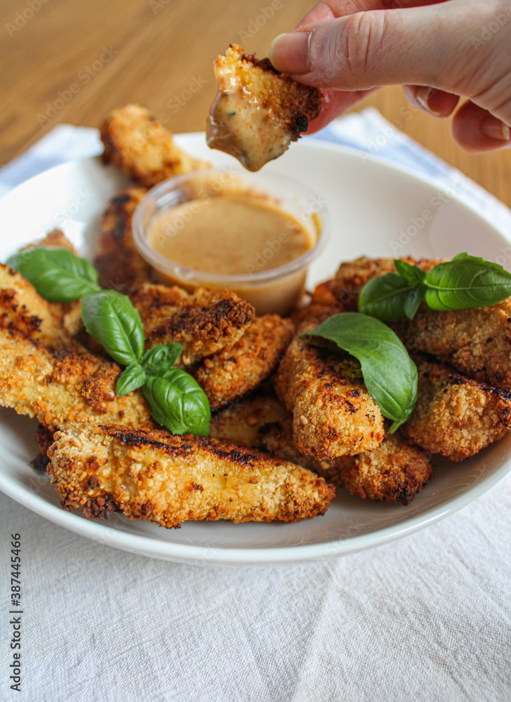 Hand holding chicken finger and dipping into the sauce. A plate of crunchy oven-baked chicken fingers with honey mustard sauce. 