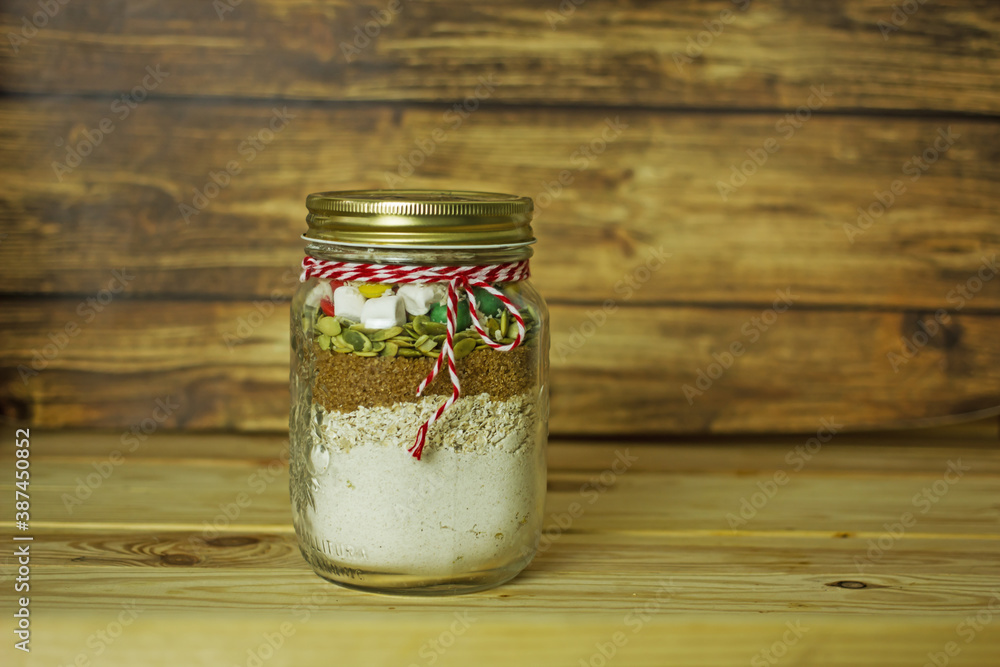 Christmas cookie mix in a jar. Flour, oatmeal, cane sugar, pumpkin seeds, marshmallows, colored drops in masson jar with a red rope on a wooden table. Christmas home cooking edible gift concept Christmas cookie mix in a jar. Flour, oatmeal, cane sugar, pumpkin seeds, marshmallows, colored drops in masson jar with a red rope on a wooden table. Christmas home cooking edible gift concept