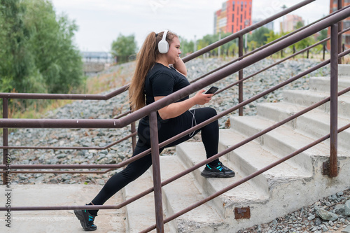 A beautiful fat girl in headphones is engaged in fitness on the sports ground and takes a selfie. Young woman lunges and is photographed on a smartphone outdoors on a warm summer day.