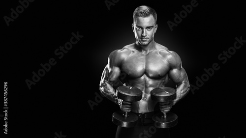 Black and white vertical photo of perfect fit athletic guy workout. Handsome power athletic man with a naked torso in training pumping up muscles with dumbbells in a gym. Fitness muscular body
