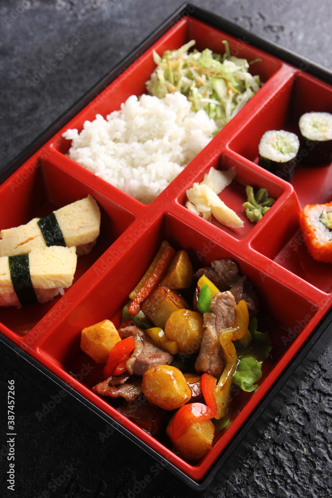 Japanese cuisine. Business lunch in a box: rolls, sushi, rice with vegetables and stew on a black table