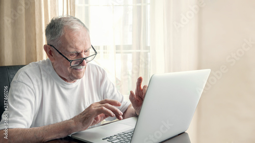 Older man at home in front of a laptop. A gray-haired old man with glasses works at home in front of a laptop. The concept of communication of old people.
