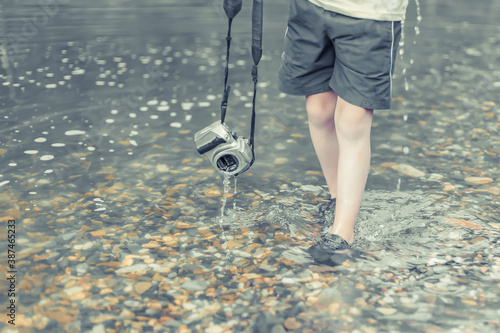 A schoolboy stands in the river on outdooor and destroys a mirrorless photo camera. The concept of obsolescence of technology. Photographers day, humor. Soft light. © Anna Shnaider