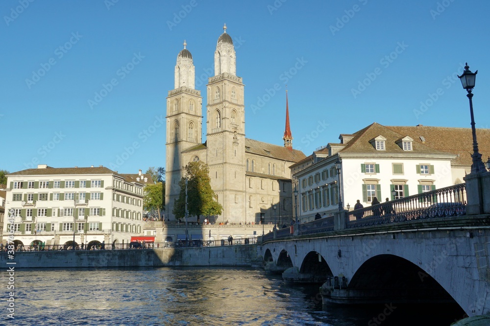 Zurich / Switzerland - 10 03 2020: Quay Bridge in Zurich in lateral view with Grossmunster cathedral on the Limmat River bank on the background surrounded by historical buildings enlighten by sun. 