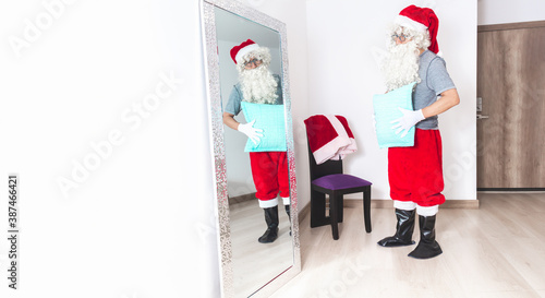 Man dressing up as Santa Claus, putting a pillow on his belly to look fatter, looking at himself in the mirror photo