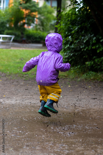 Fototapeta Naklejka Na Ścianę i Meble -  Child from behind jumping into a puddle of mud during a rainy and wet summer day at kindergarten