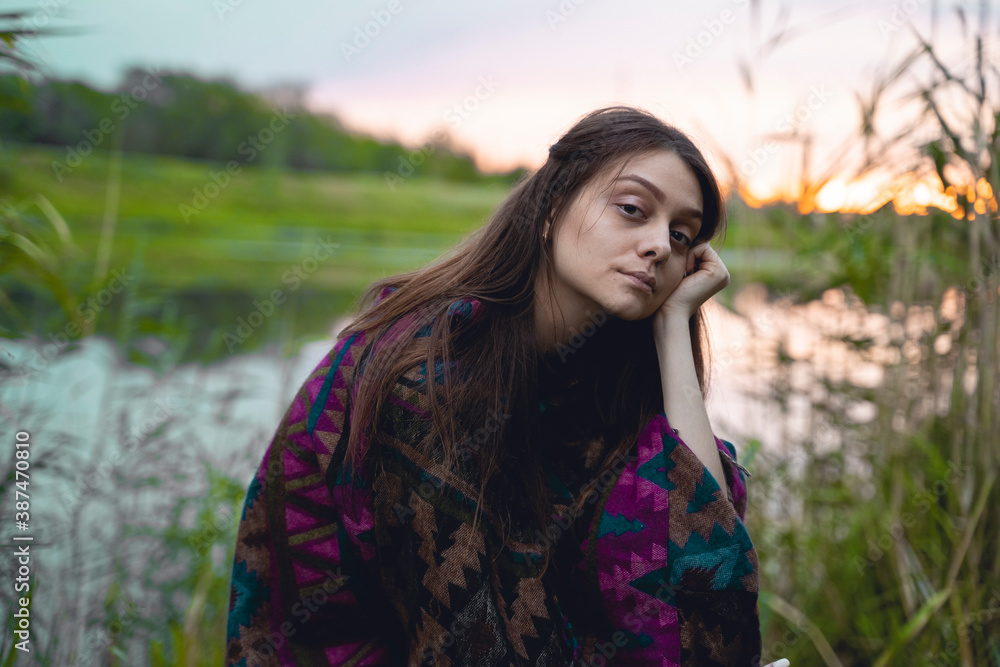 A young woman in an indian poncho sits in green grass on a sunset background