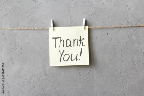 Note Thank you! is written on a yellow sticker that hangs with a clothespins on a grey background