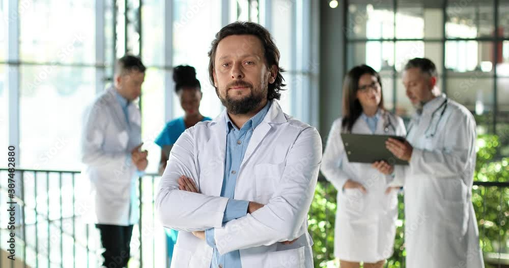 Portrait of Caucasian handsome man physician looking at camera and standing in clinic. Indoors. Male doctor in hospital. Mixed-races docs talking on background. Medic in white gown. Portrait of Caucasian handsome man physician looking at camera and standing in clinic. Indoors. Male doctor in hospital. Mixed-races docs talking on background. Medic in white gown.