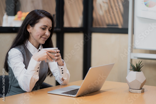 writes new posts for social networks and their subscribers. European-looking brunette sitting in a coworking office, looking at the camera. confident female blogger working on a laptop