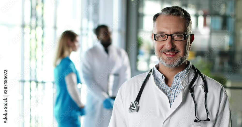Portrait shot of Caucasian senior man physician in white gown, stethoscope and glasses looking at camera and smiling in clinic. Handsome male doctor in hospital. Doctors on background Medicine concept Portrait shot of Caucasian senior man physician in white gown, stethoscope and glasses looking at camera and smiling in clinic. Handsome male doctor in hospital. Doctors on background Medicine concept