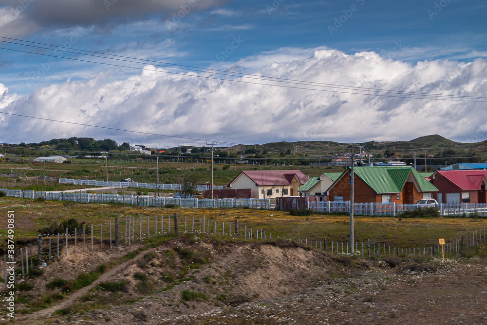 Punta Arenas, Chile - December 12, 2008: Houses in green meadows in rural area outside city center with white belt of huge cloudscape under blue sky.