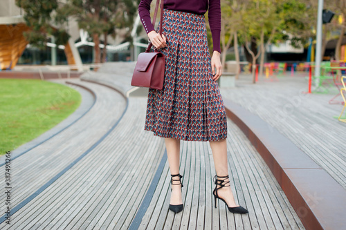 Fashionable young woman wearing pleated midi skirt, sweater, black high heel shoes and holding burgundy handbag in hand on the city street. Street style.