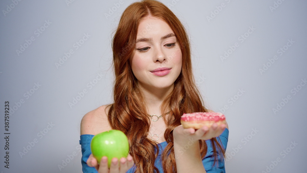 Doubt female person making a decision in studio. Girl choosing doughnut indoors Doubt female person making a decision in studio. Girl choosing doughnut indoors