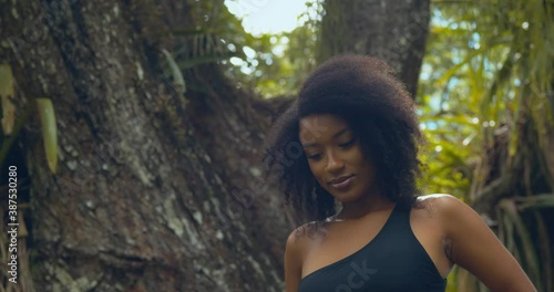 With rain drops falling in the background a natural hair young lady stands on the large tree trunk of a silk cotton tree photo