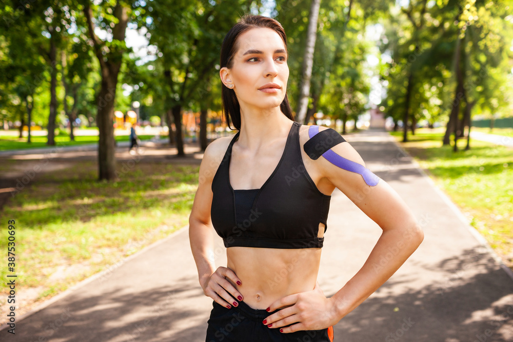 Portrait of attractive muscular brunette woman wearing black sports outfit, looking at camera. Young smiling female athlete posing with hands on waist, colorful kinesiotaping on body. Portrait of attractive muscular brunette woman wearing black sports outfit, looking at camera. Young smiling female athlete posing with hands on waist, colorful kinesiotaping on body.