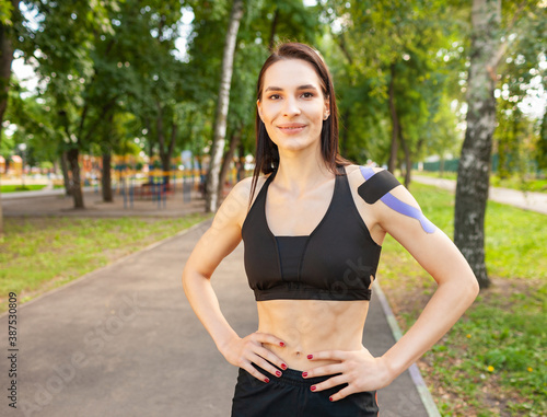 Portrait of attractive muscular brunette woman wearing black sports outfit, looking at camera. Young smiling female athlete posing with hands on waist, colorful kinesiotaping on body. © YouraPechkin
