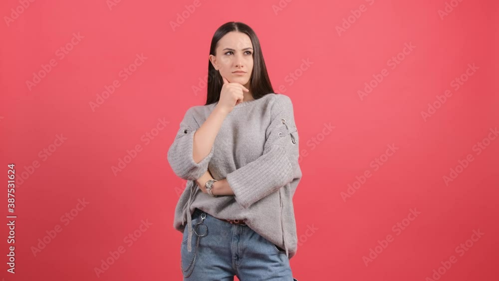 Serious young woman sits with her hand next to face and looks aside as she thinks about important things, isolated over red background. Serious young woman sits with her hand next to face and looks aside as she thinks about important things, isolated over red background.