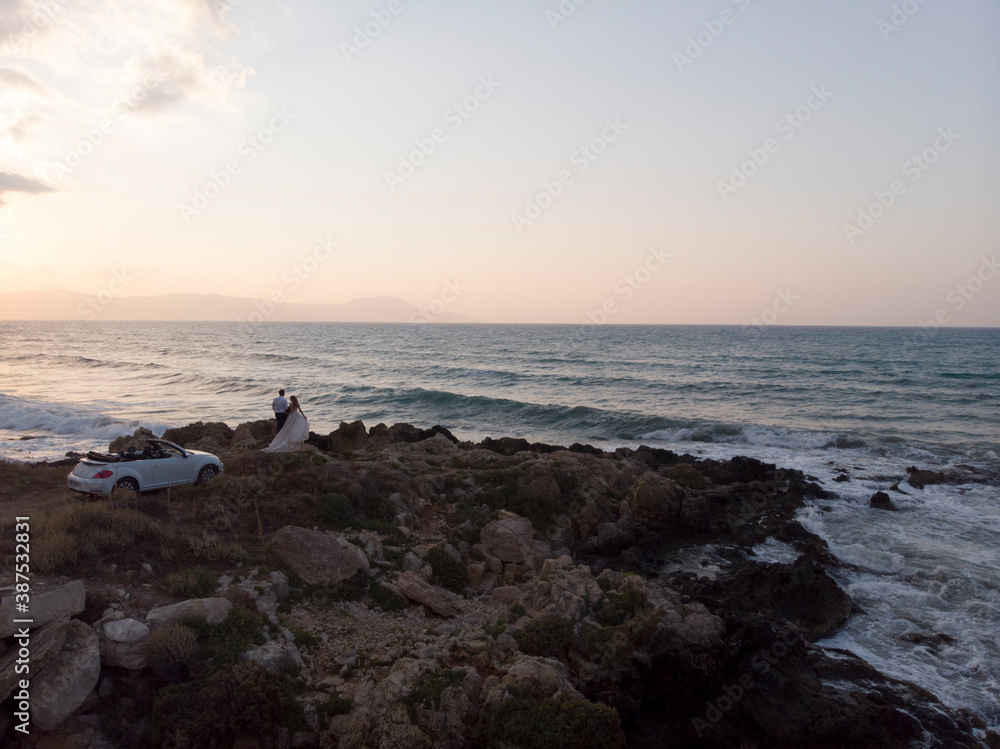 Bride in white fluffy dress and groom in tuxedo hugging on rocky coast, shore near the sea at sunset, white cabriolet car stand near, Crete, Greece. Wedding love story. Aerial.