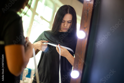 Young woman designer measuring her breast with tape measure on front of mirror at workshop