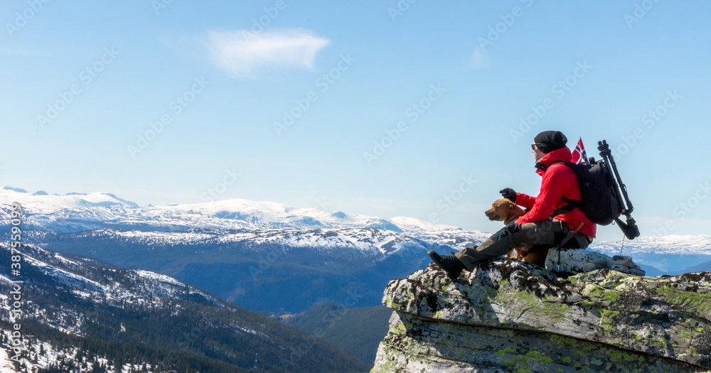 Man and dog standing on steep hillside and looking towards the snowy peaks of Jotunheimen in Norway. Staffordshire bullterrier, hiking, trekking, lifestyle, rondane and Norway concept. Man and dog standing on steep hillside and looking towards the snowy peaks of Jotunheimen in Norway. Staffordshire bullterrier, hiking, trekking, lifestyle, rondane and Norway concept.