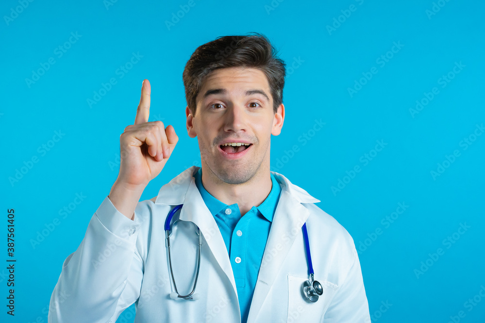 Portrait of doctor man in medical coat having idea moment pointing finger up on blue studio background. Smiling happy doc showing eureka gesture. Portrait of doctor man in medical coat having idea moment pointing finger up on blue studio background. Smiling happy doc showing eureka gesture.