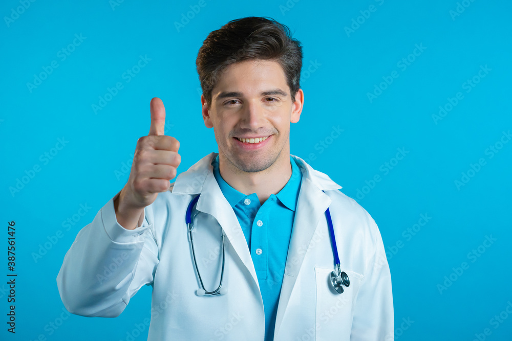 Doctor showing thumb up sign over blue background. Positive young man in medical coat smiles to camera. Winner. Success. Body language. Doctor showing thumb up sign over blue background. Positive young man in medical coat smiles to camera. Winner. Success. Body language.