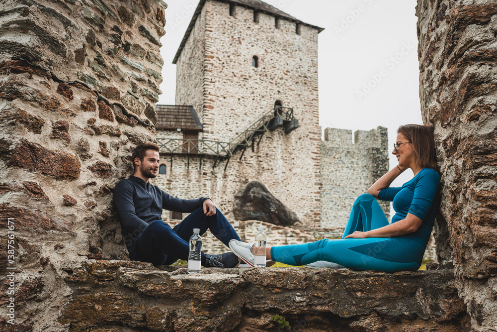 couple sitting on a stone wall in front of an old fortress and taking a break from running in forest