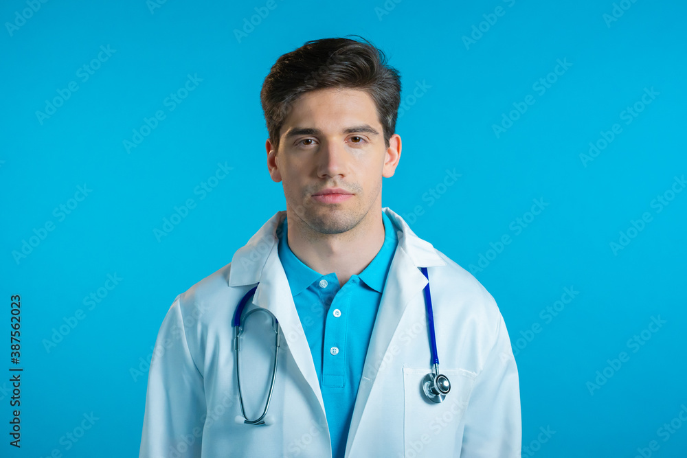 Handsome doctor man in professional white coat on blue studio background. Serious doc looking to camera Handsome doctor man in professional white coat on blue studio background. Serious doc looking to camera