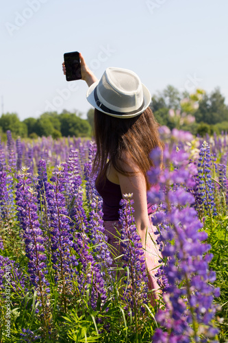 Beautiful girl takes a selfie on the field with lupins, shooting from the back