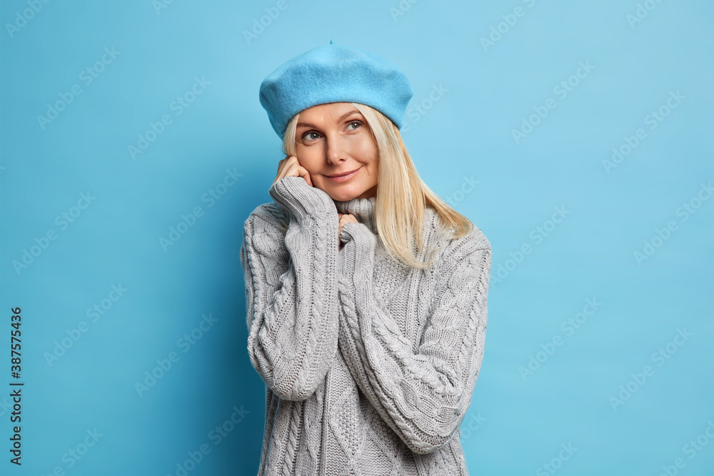 Photo of good looking middle aged woman has dreamy pensive expression wears cozy winter grey sweater and blue beret feels optimistic in good expectations going to go out during rainy autumn day Photo of good looking middle aged woman has dreamy pensive expression wears cozy winter grey sweater and blue beret feels optimistic in good expectations going to go out during rainy autumn day