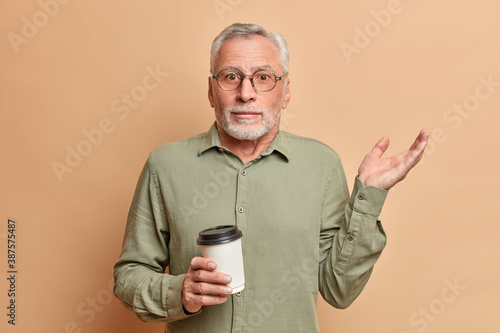 Confused bearded old man shrugs shoulders and raises hand being clueless and doubtful to give answer has coffee break dressed in shirt poses against beige background. Human reaction perception © wayhome.studio 