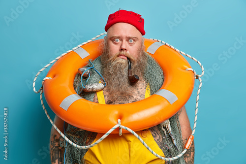Stunned bearded man stares bugged eyes poses with fishing net lifebuoy and smokes pipe spends time on boat during sea voyage isolated over blue background. Surprised fisher or seafarer indoor photo