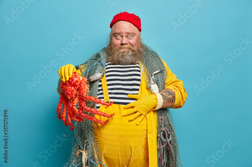 Cheerful bearded fisherman keeps hand on belly happy to catch big octopus enjoys marine adventure and voyage looks surprisingly at camera poses indoor against blue background. Middle aged sailor