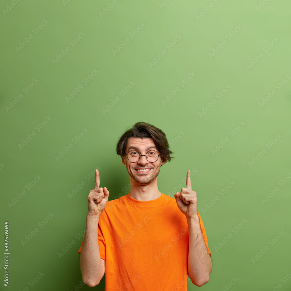 Studio shot of happy unshaven European man with positive expression points upwards on empty space demonstrates advertising content against green background suggests big sale discounts or good offer Studio shot of happy unshaven European man with positive expression points upwards on empty space demonstrates advertising content against green background suggests big sale discounts or good offer
