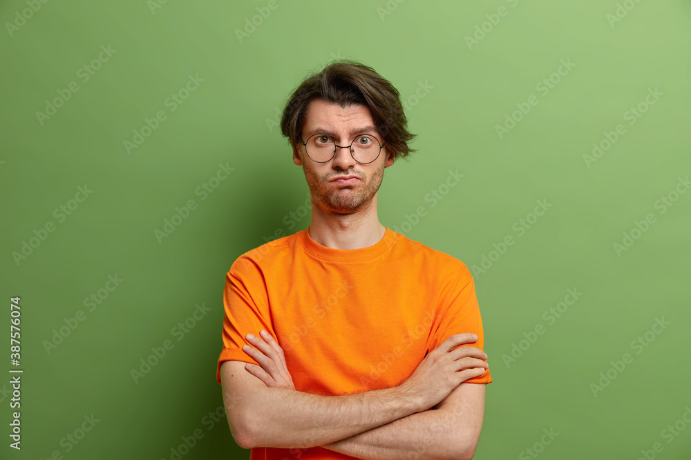 Serious handsome man in spectacles with trendy hairstyle stands in assertive pose keeps arms folded listens attentively interlocutor dressed in casual t shirt stands indoor against green background Serious handsome man in spectacles with trendy hairstyle stands in assertive pose keeps arms folded listens attentively interlocutor dressed in casual t shirt stands indoor against green background