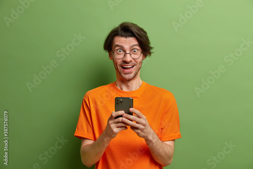 Cheerful European hipster guy plays video game on smartphone uses high speed internet surfs social networks dressed in casual orange t shirt poses against vivid green background. Technology concept