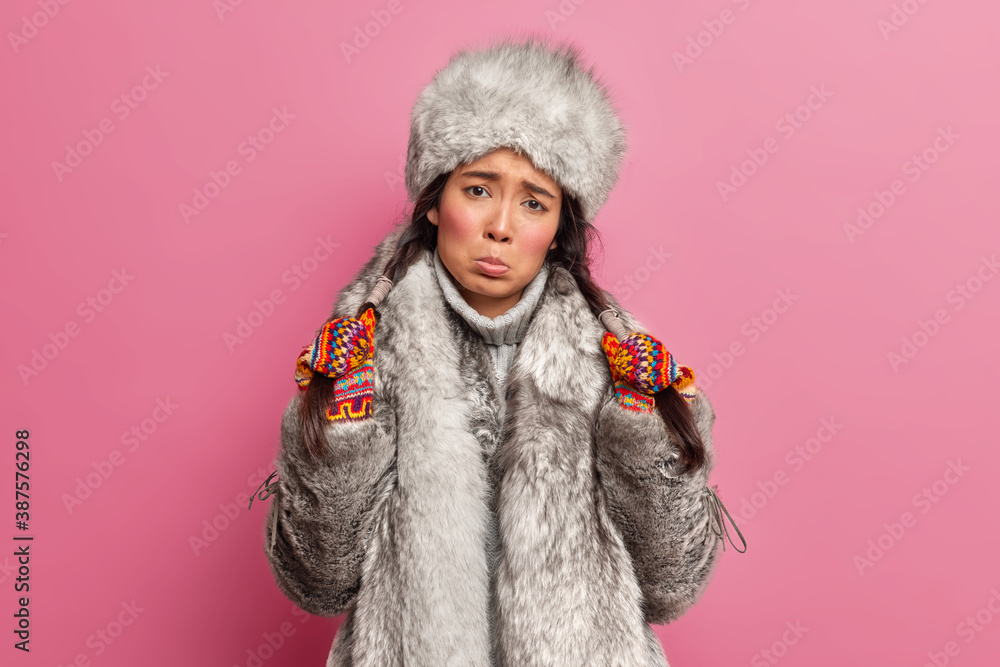 Upset Eskimo woman looks with gloomy expression at camera holds two pigtails wears warm winter clothes being resident of Alaska or Chukotka lives on north pole stands against rosy background. Upset Eskimo woman looks with gloomy expression at camera holds two pigtails wears warm winter clothes being resident of Alaska or Chukotka lives on north pole stands against rosy background.