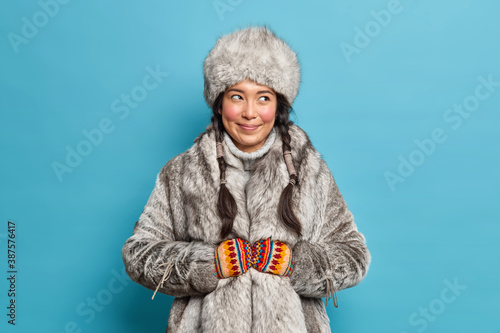Pleased smiling eskimos woman dressed in traditional clothing adapted to extreme cold lives on nothern pole isolated over blue background. Female inhabitant of Alaska wears fut coat and hat.
