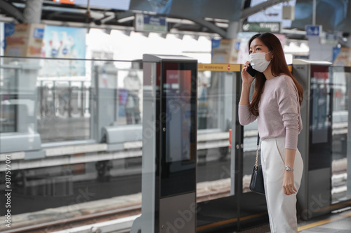 Asian business woman in casual clothes wearing face mask talking on mobile phone. She is waiting for the train to go to work on the platform station. New normal lifestyle in city concept.