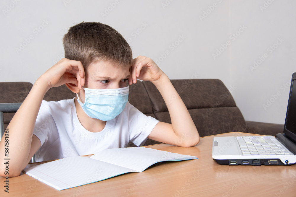 A child in a protective mask on his face looks displeasedly at a laptop, a boy learns remotely online at home