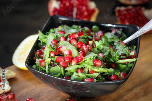 Tabbouleh with pomegranate. Green salad of parsley and herring with large pomegranate grains, in a black plate on a dark background and a wooden board.