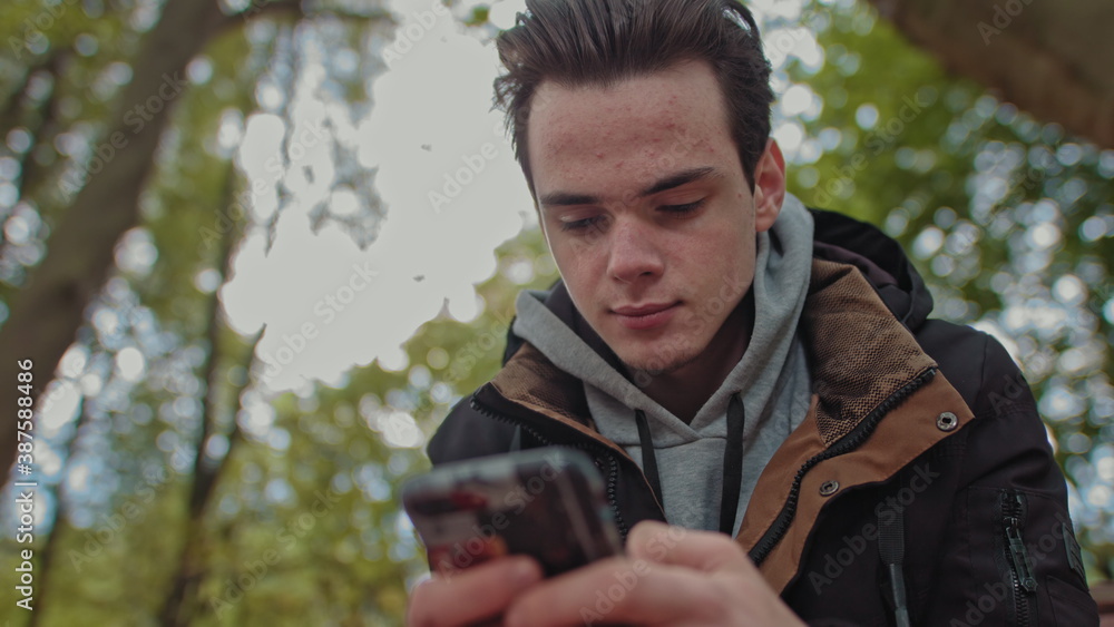 Teenager Looks at the phone, dials a message on a touch screen phone. Shooting close up of a smile on his face. The guy is sitting in the park on a bench. Corresponds with friends Teenager Looks at the phone, dials a message on a touch screen phone. Shooting close up of a smile on his face. The guy is sitting in the park on a bench. Corresponds with friends