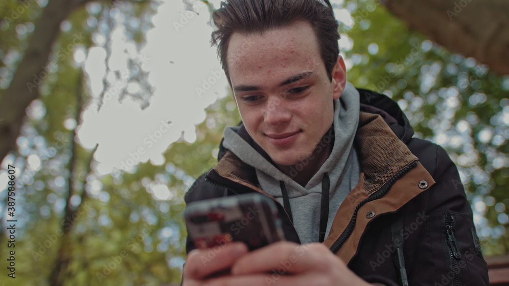 Teenager Looks at the phone, dials a message on a touch screen phone. Shooting close up of a smile on his face. The guy is sitting in the park on a bench. Corresponds with friends